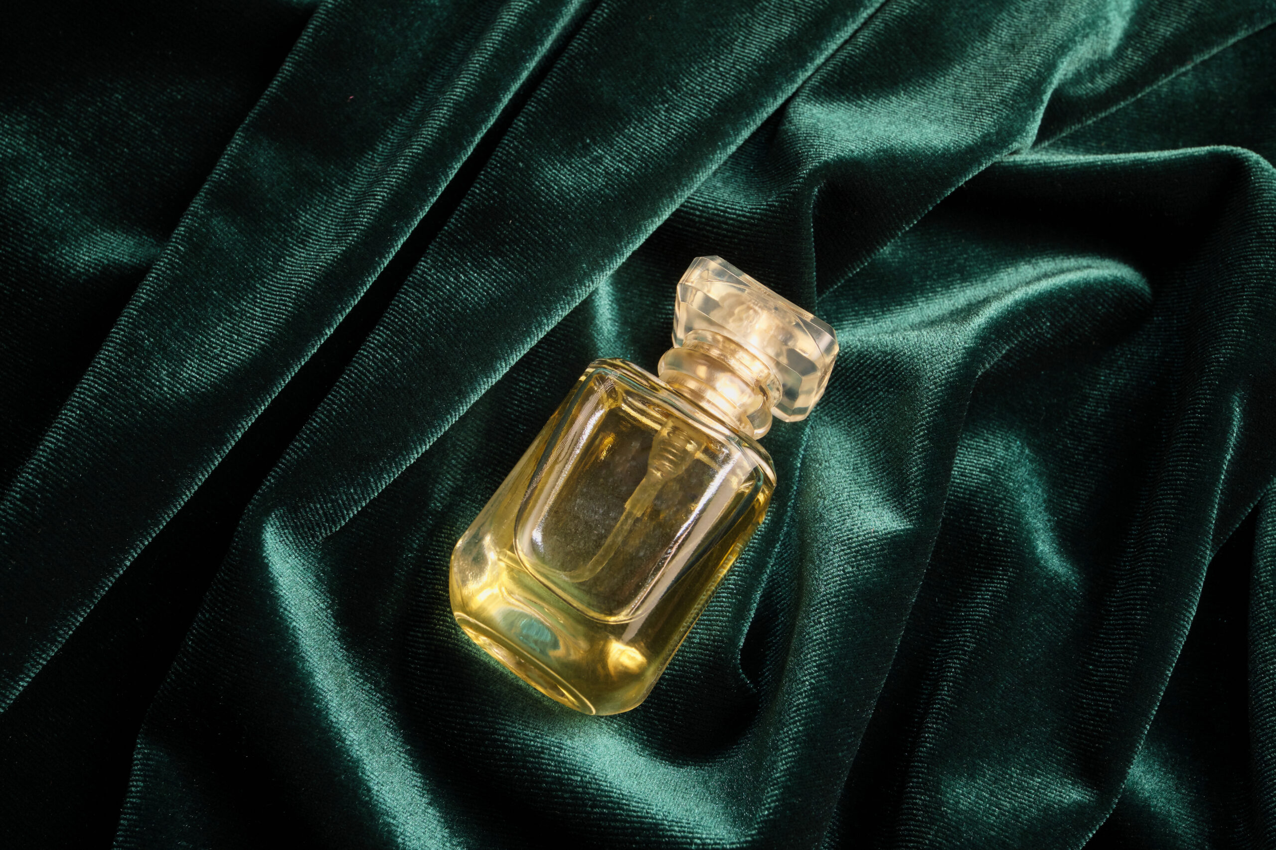 Frasco de perfume de cristal sobre terciopelo verde, con reflejos cálidos y textura visible en las sombras.